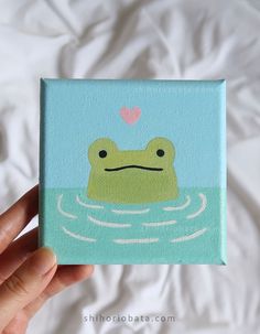 a hand holding up a small square painting with a frog on it