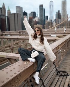 a woman in white sweater and black pants sitting on top of a bridge with her arms up