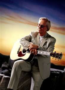 a man sitting on top of a rock with a guitar in his hand and the sun setting behind him