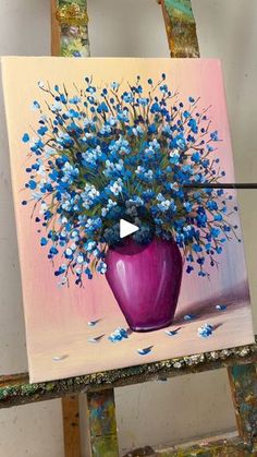 a purple vase with blue flowers in it on a easel next to paintbrushes