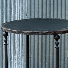 a round table with two black legs and a metal top, against a corrugated wall