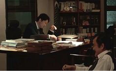 two men sitting at a desk with books and papers in front of them, one is talking on the phone