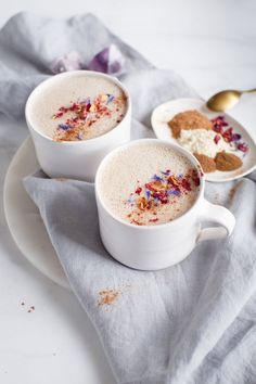 two white cups filled with hot chocolate and sprinkles on top of a plate