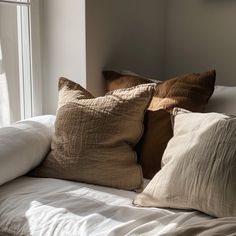 a bed topped with lots of pillows next to a window covered in white linens