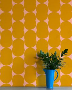 a blue vase filled with green plants on top of a wooden table next to a yellow wall