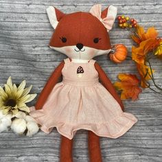 a stuffed fox wearing a pink dress next to flowers and pumpkins on a wooden surface