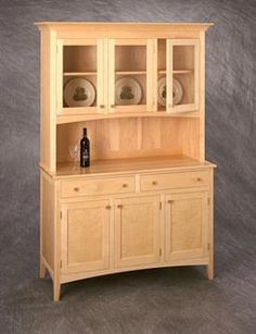 a wooden hutch with glass doors and plates on the top, in front of a gray background