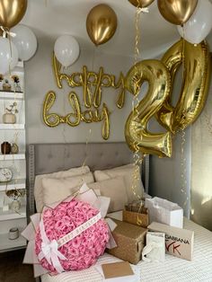 balloons and presents are on the bed in this room with gold foil letters, white sheets, and pink roses