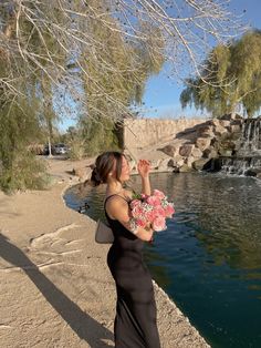 a woman in a long black dress is holding flowers near a pond with waterfall behind her