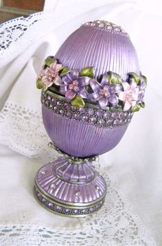 an egg decorated with purple flowers on top of a white table cloth next to a lace doily