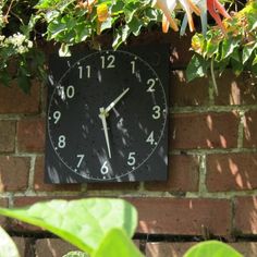 a black clock mounted to the side of a brick wall surrounded by green plants and flowers