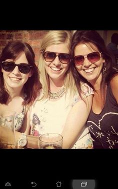 three women are posing for the camera with drinks in front of them and brick wall behind them