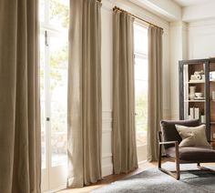 a living room filled with furniture and windows covered in beige drapes, next to a bookcase