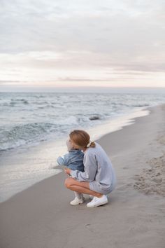 a woman kneeling down holding a baby on top of a beach next to the ocean