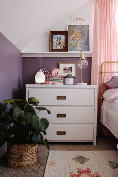 a white dresser sitting next to a bed in a room with pink curtains and pictures on the wall