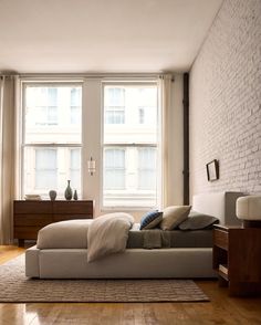 a bed sitting in the middle of a living room next to two large windows and a wooden floor