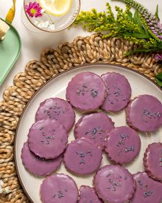 purple frosted cookies on a plate next to some flowers and lemon wedges in a basket