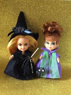 two dolls sitting next to each other in front of a gold glitter background, one holding a broom and the other wearing a witch costume