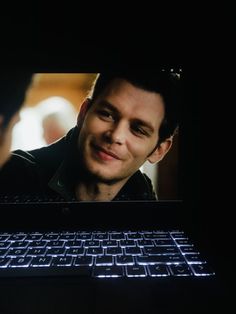 a laptop computer with a man on the screen and another person in the background looking at it