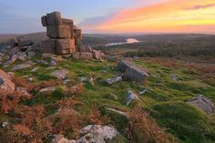 the sun is setting over some rocks and grass on top of a hill with water in the distance