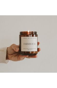 a hand holding a jar of farm house candle in front of a white background with the label on it