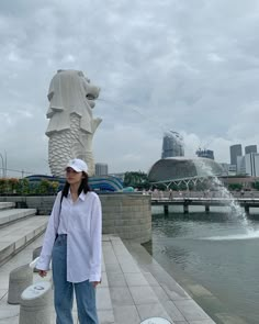 a woman standing in front of a fountain with a large statue behind her and buildings in the background