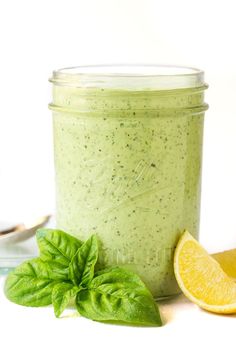 a jar filled with green smoothie next to a lemon slice and basil leaf on a white surface