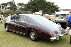 an old car is parked on the grass near other antique cars and people standing around