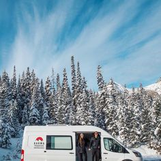 two people standing in the open door of a van on a snowy mountain side with trees behind them