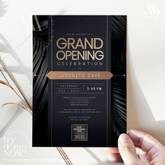 a hand holding up a black and gold grand opening card with palm leaves in the background