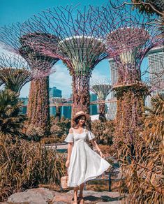 a woman in a white dress and straw hat walking through the gardens by the bay