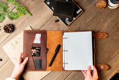 a person's hand holding a pen and notebook on top of a wooden table