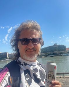 a man holding up a can of beer in front of a body of water with buildings in the background