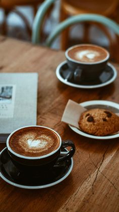 two cups of cappuccino and a cookie are sitting on a wooden table