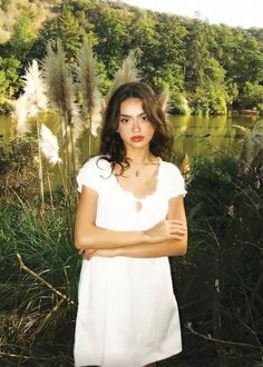 a woman standing in front of a lake with her arms crossed and looking at the camera