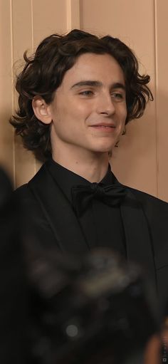 a young man in a black shirt and bow tie smiles at the camera as he stands next to a wall