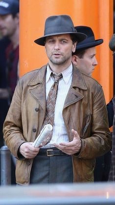 a man wearing a brown jacket and tie standing next to another man in a hat