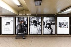 a man walking past posters on the wall in a subway station with people standing next to them