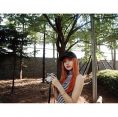 a woman with red hair is holding a camera and posing in front of a tree