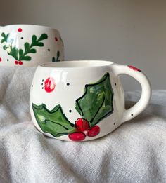 two ceramic mugs with holly designs on them are sitting on a white cloth covered surface