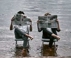 two people sitting on chairs in the water with newspapers covering their faces and reading them