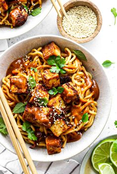 two white bowls filled with noodles and tofu on top of a table next to chopsticks