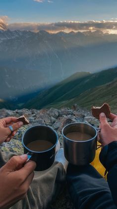 two people holding cups of coffee on top of a mountain with mountains in the background