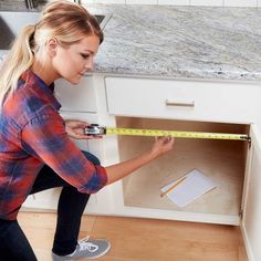 a woman measuring the width of a kitchen cabinet in front of a counter top with a tape on it