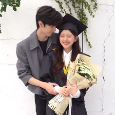 a young man and woman in graduation gowns holding bouquets, smiling at each other