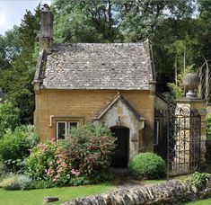 an old stone house surrounded by greenery and flowers with a gate in the foreground