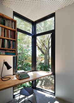 a desk with a keyboard, lamp and bookshelf in front of large windows