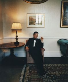 a man in a tuxedo sitting on a chair next to a table and lamp