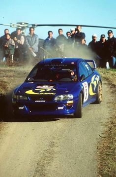 a blue car driving down a dirt road next to a group of people and a helicopter