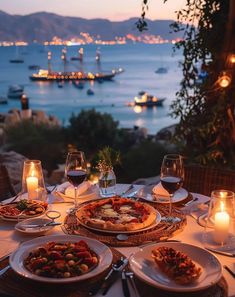 an outdoor table with plates and glasses of wine on it, overlooking the water at night